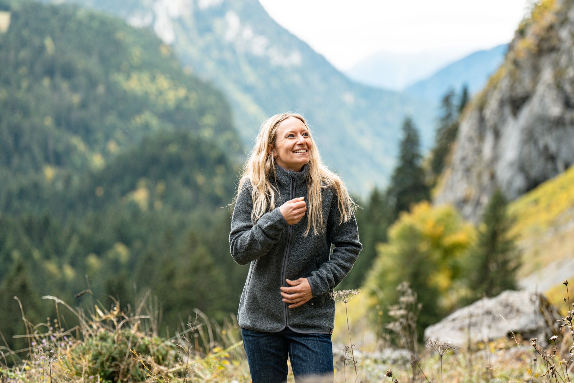 Une femme souriante en veste polaire en Merino avec une montagne boisée en arrière-plan